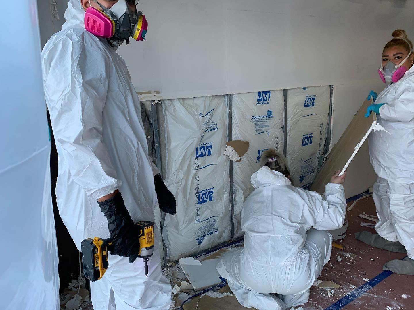 Three individuals in protective suits, masks, and gloves are working on removing drywall with exposed insulation behind it. One person holds a power drill while another uses a tool to pry off the drywall, all part of an extensive water damage remediation process.